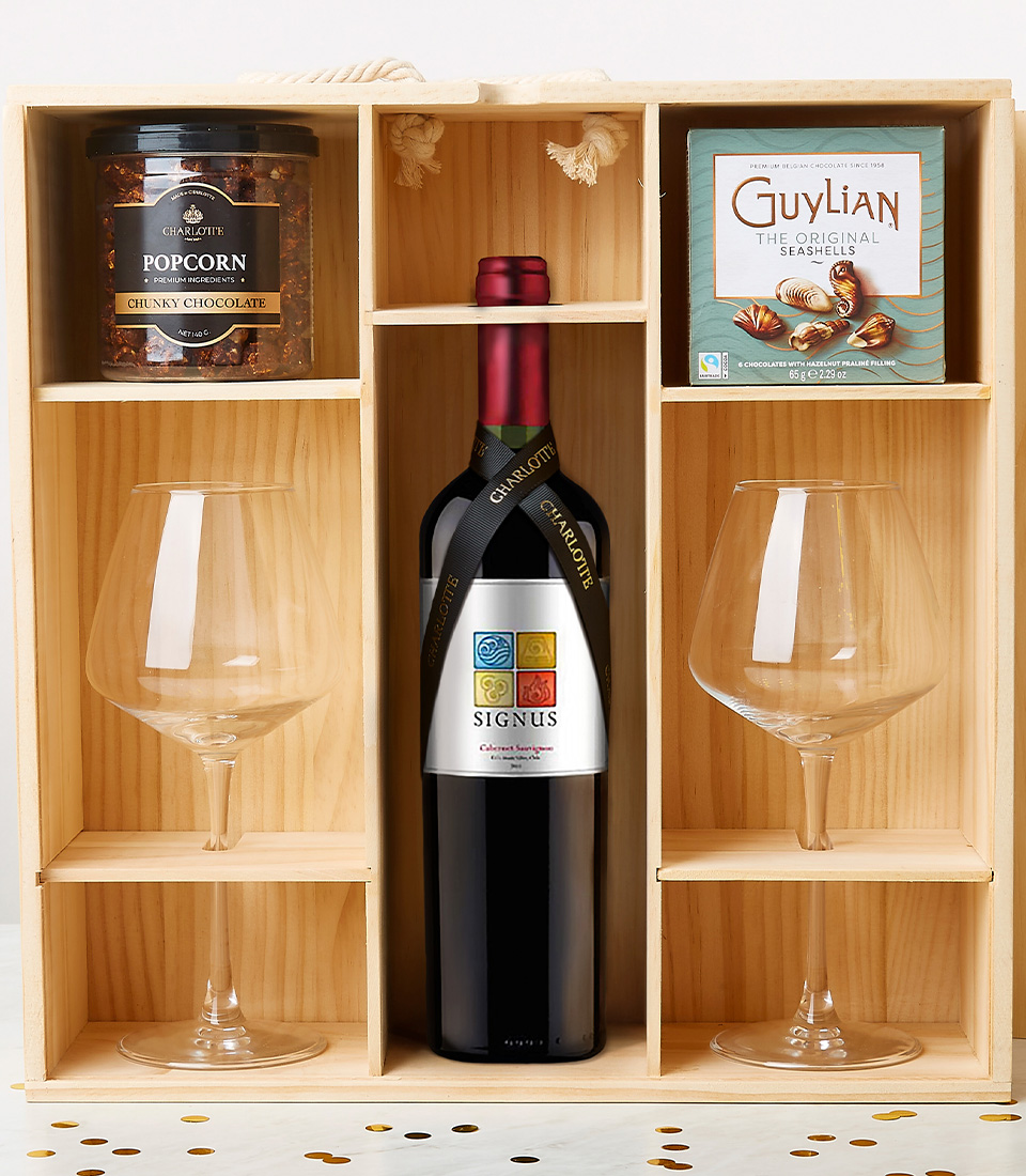 A luxurious wooden gift box from Basketeer elegantly showcases a bottle of exquisite Tempus Two wine, flanked by two crystal wine glasses. Nestled above the bottle, a jar of decadent organic chocolate-coated popcorn sits alongside a sophisticated box of Guylian Belgian chocolates. Delicate decorative confetti is artfully scattered below, adding an extra touch of celebration and opulence to the scene. The prestigious Basketeer logo graces a ribbon and box within the arrangement, seamlessly tying together this invitation to indulge in refined elegance.