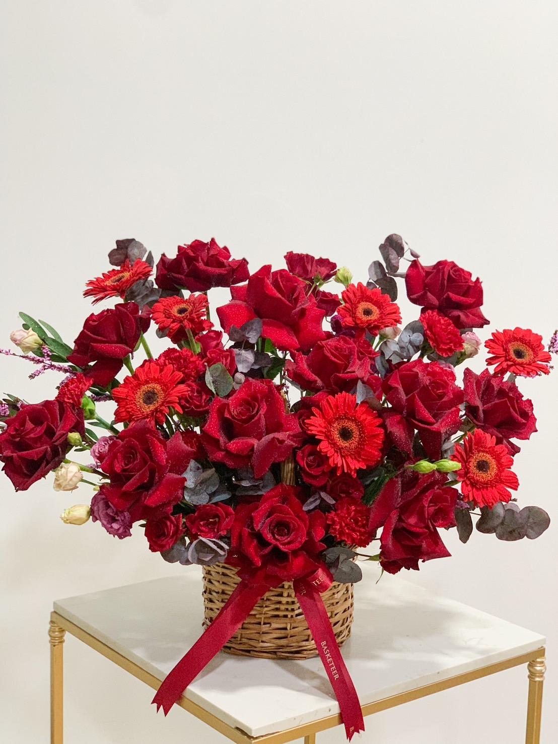 A basket filled with a vibrant arrangement of red roses and red gerbera daisies, interspersed with green eucalyptus leaves. The basket, adorned with a red bow, sits on a small rectangular table with a white top and golden legs. The background is a plain white wall.