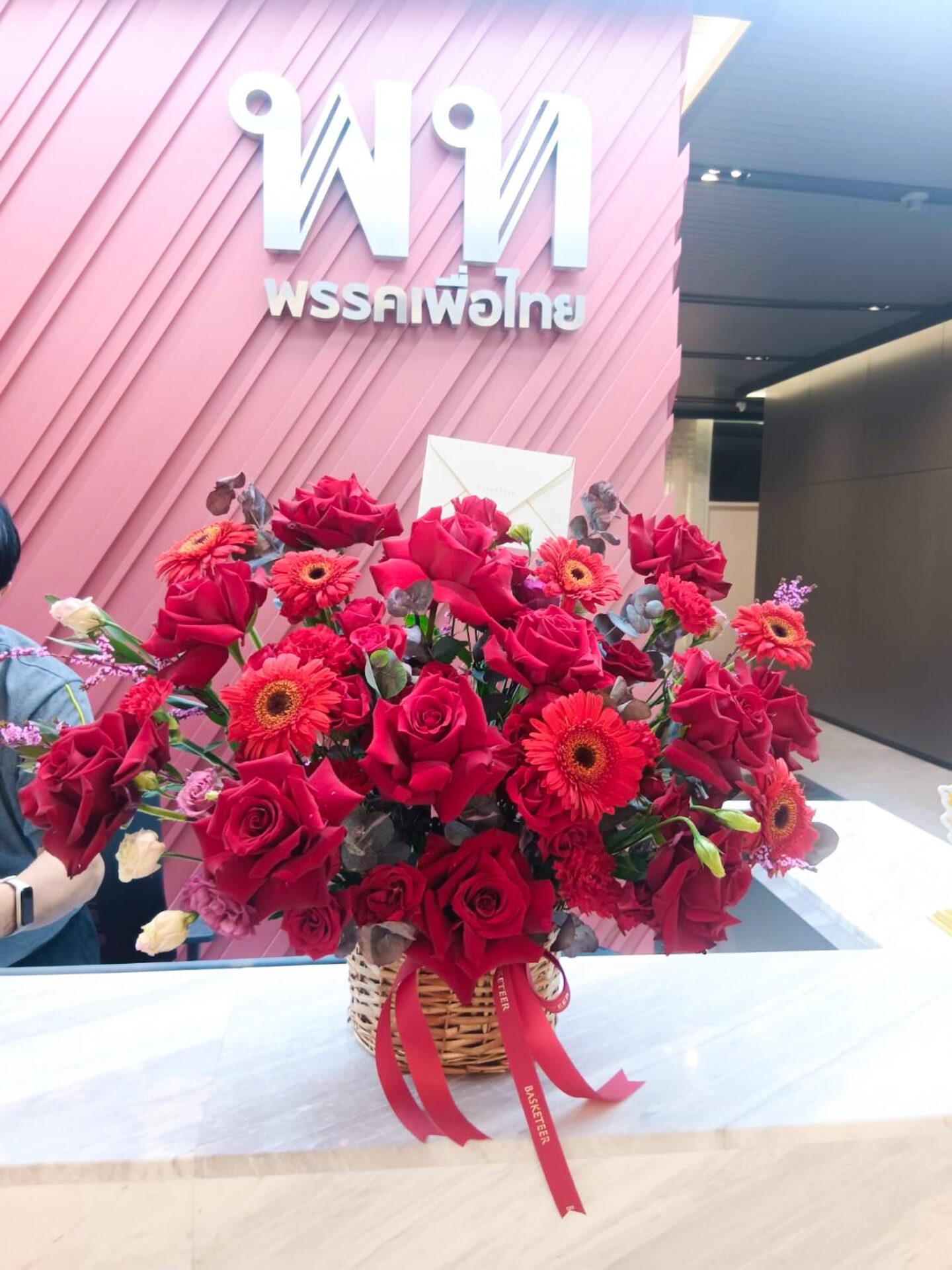 A basket of vibrant red roses and gerbera daisies adorned with a red ribbon sits on a white counter. Behind it is a wall with a pink geometric design and Thai text. A person is partially visible to the left.