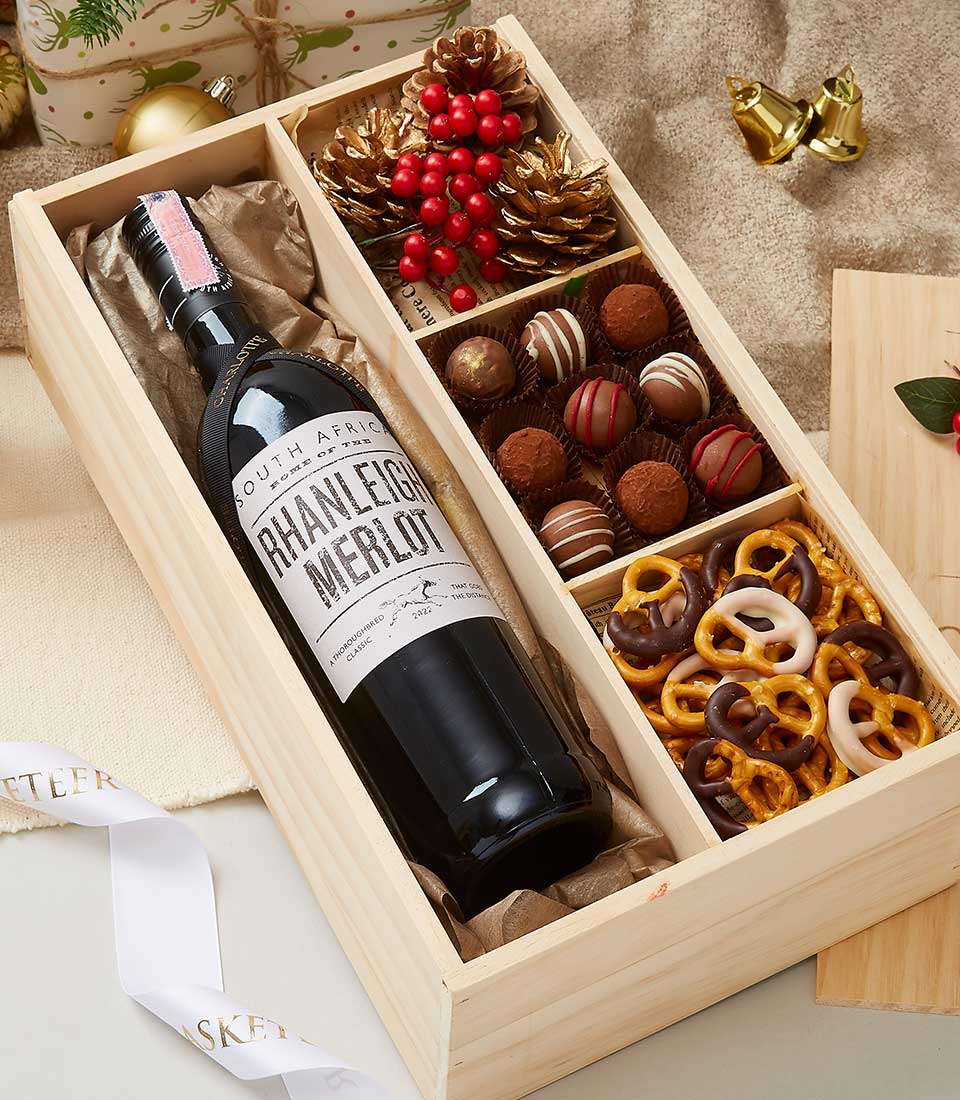 A luxurious wooden gift box exquisitely houses a bottle of South African Rhanleigh Merlot on the left, accompanied by an opulent selection of chocolates, decadent truffles, and indulgent chocolate-covered pretzels on the right. Surrounding this elegant presentation are festive touches—pinecones, vibrant red berries, and glistening gold bells—setting a warm and inviting holiday scene. A tasteful ribbon or box adorned with the distinctive Basketeer logo adds an extra touch of sophistication to this lavish offering.
