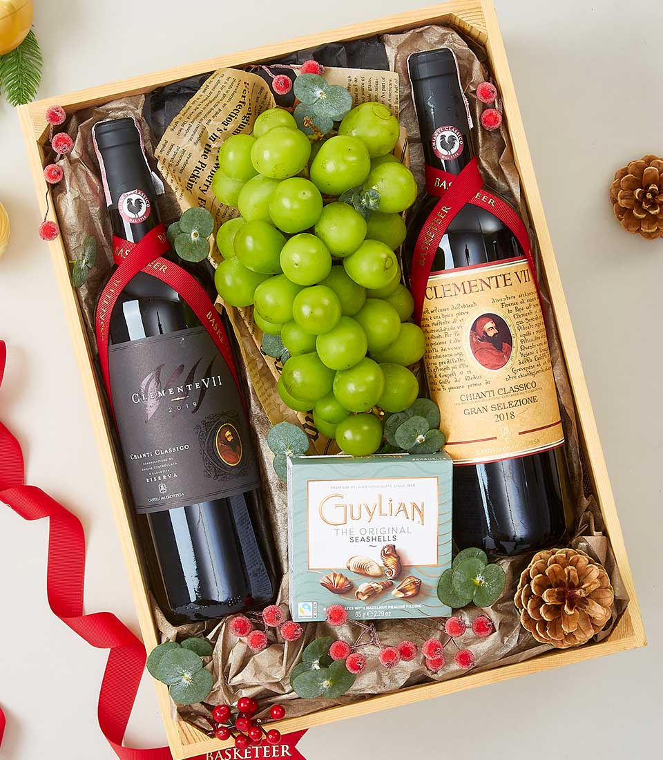 A lavish wooden gift box from Basketeer showcasing two exquisite bottles of red wine nestled beside a succulent bunch of green grapes. Accompanying these delights is an elegant box of Guylian chocolates, perfect for indulging in luxury. The presentation is enhanced with artful touches: decorative pine cones, a rich red ribbon adorned with the Basketeer logo, and intricate accents of vibrant red berries and shimmering golden leaves. This opulent ensemble invites moments of pure bliss and refined taste.