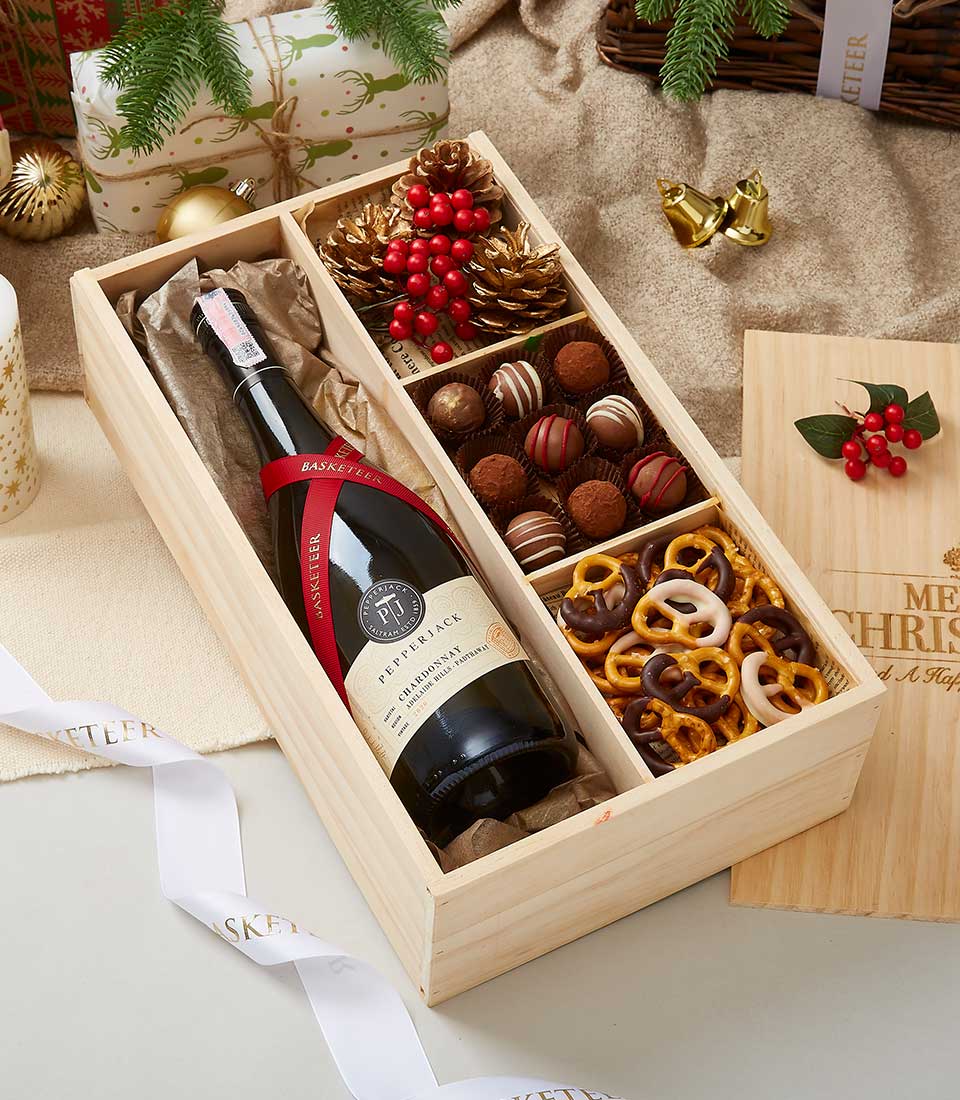 A wooden gift box containing a bottle of red wine and an assortment of chocolates and pretzels is displayed. The box is surrounded by festive holiday decorations, including pinecones, berries, and wrapped gifts, suggesting a Christmas or holiday theme.