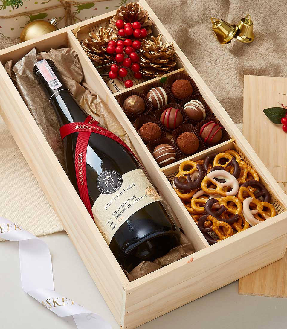 A beautifully handcrafted wooden gift box featuring the exquisite Pepperjack Barossa Shiraz wine on the left, accompanied by a sumptuous selection of chocolates and decadent chocolate-covered pretzels on the right. Nestled within the arrangement are elegant pinecones and festive red berries for a seasonal touch. The background showcases luxurious holiday-themed bell decorations, while an opulent ribbon adorned with the Basketeer logo adds a sophisticated flair to this indulgent gift experience.