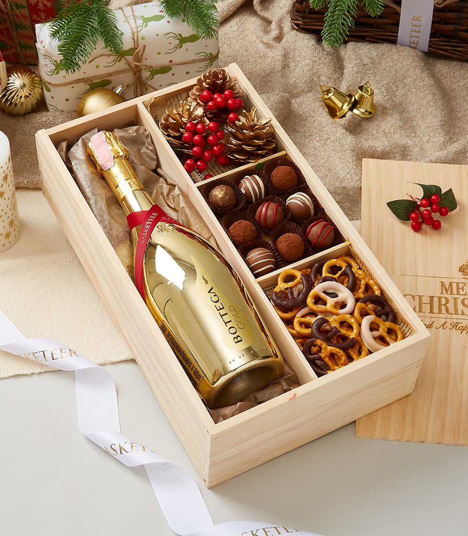 A lavish Basketeer wooden gift box elegantly showcases a bottle of Bottega Gold Prosecco in its gleaming splendor, accompanied by meticulously arranged compartments brimming with an exquisite variety of assorted chocolates and pretzels. The scene is adorned with festive holiday decorations—rustic pinecones, vibrant red berries, and beautifully wrapped gifts, creating an inviting atmosphere of celebration. A luxurious ribbon proudly bearing the Basketeer logo completes this opulent presentation, making it a perfect indulgence for any special occasion.