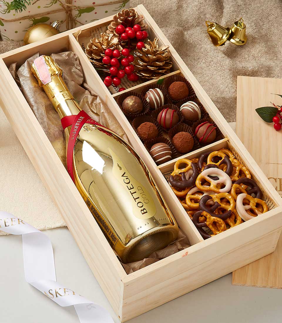 A luxurious wooden gift box from Basketeer featuring a bottle of Bottega Gold prosecco, an assortment of decadent chocolate truffles, and artisanal chocolate-covered pretzels. The arrangement is adorned with festive decorations including elegant pinecones and vibrant red berries, resting gracefully on a plush fabric surface. Nearby lies a delicate white ribbon and gleaming golden bells, evoking an air of opulence and celebration.