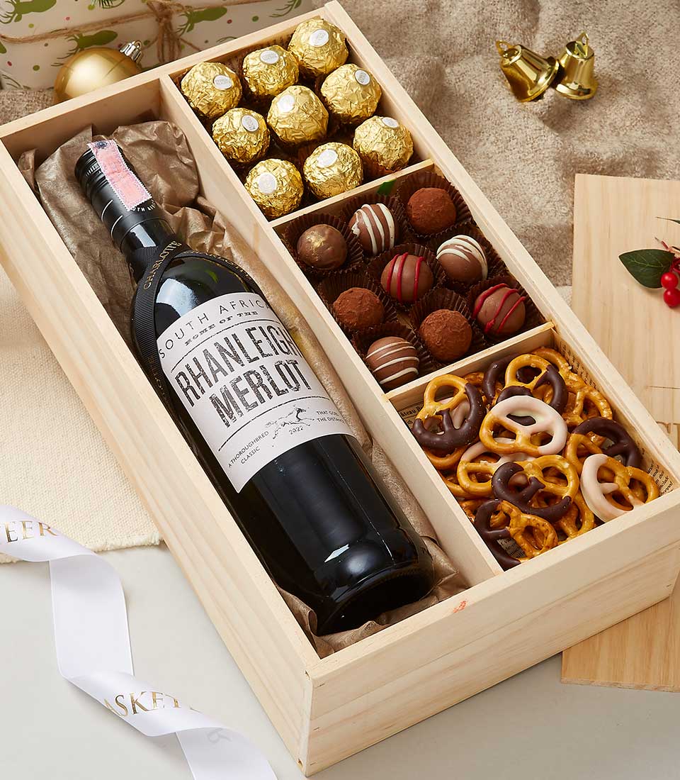 An elegant wooden gift box by Basketeer, featuring a premium bottle of South African red wine, an exquisite selection of assorted chocolates, and indulgent chocolate-covered pretzels. The luxurious box is showcased on a sumptuous beige cloth adorned with festive Christmas ornaments—gold bells and a charming wreath embellished with vibrant red berries. A ribbon graced with the Basketeer logo adds an extra touch of sophistication to this holiday treasure.