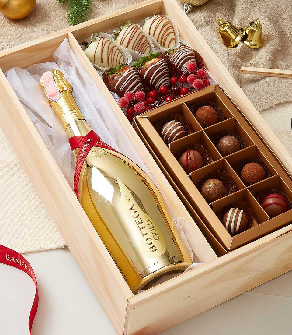 An opulent Basketeer gift box exuding luxury and sophistication. Nestled within a meticulously crafted wooden compartmentalized box, you'll find an exquisite bottle of Bottega Gold sparkling wine on the left. The scene is decadently adorned with chocolate-dipped strawberries, gourmet truffles, and a beautiful arrangement of red berries. A luxurious red ribbon elegantly embellished with the distinctive Basketeer logo completes this inviting presentation, making it the perfect indulgence for any special occasion.