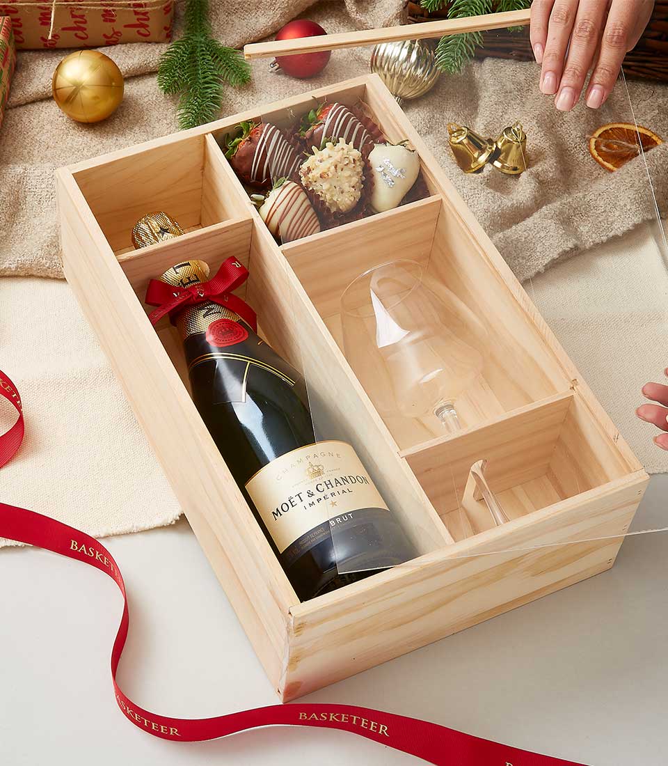 A lavish wooden gift box, elegantly partitioned, showcases a bottle of prestigious Moët & Chandon champagne, an exquisite wine glass, and a delectable selection of chocolate-covered strawberries. The scene captures a graceful hand gently placing the lid adorned with the Basketeer logo onto the box. Surrounding this opulent gift are decorative ribbons and festive ornaments that exude an air of celebration and luxury.