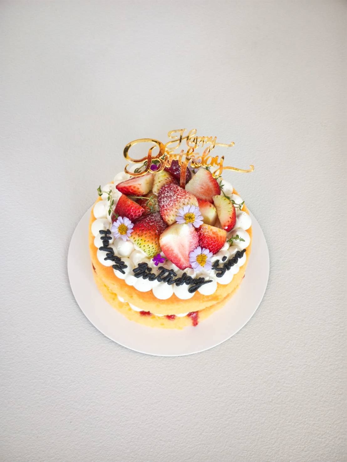 Fresh strawberry birthday cake topped with whipped cream, sliced strawberries, and small edible flowers, with a gold “Happy Birthday” topper.