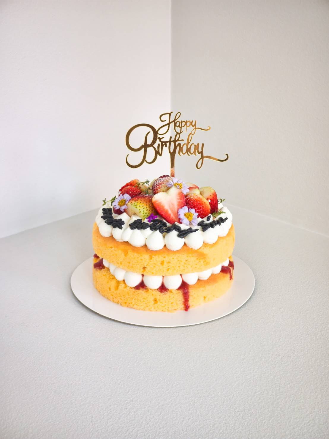 Two-layer strawberry birthday cake with whipped cream, sliced strawberries, and edible flowers, topped with a gold “Happy Birthday” sign.