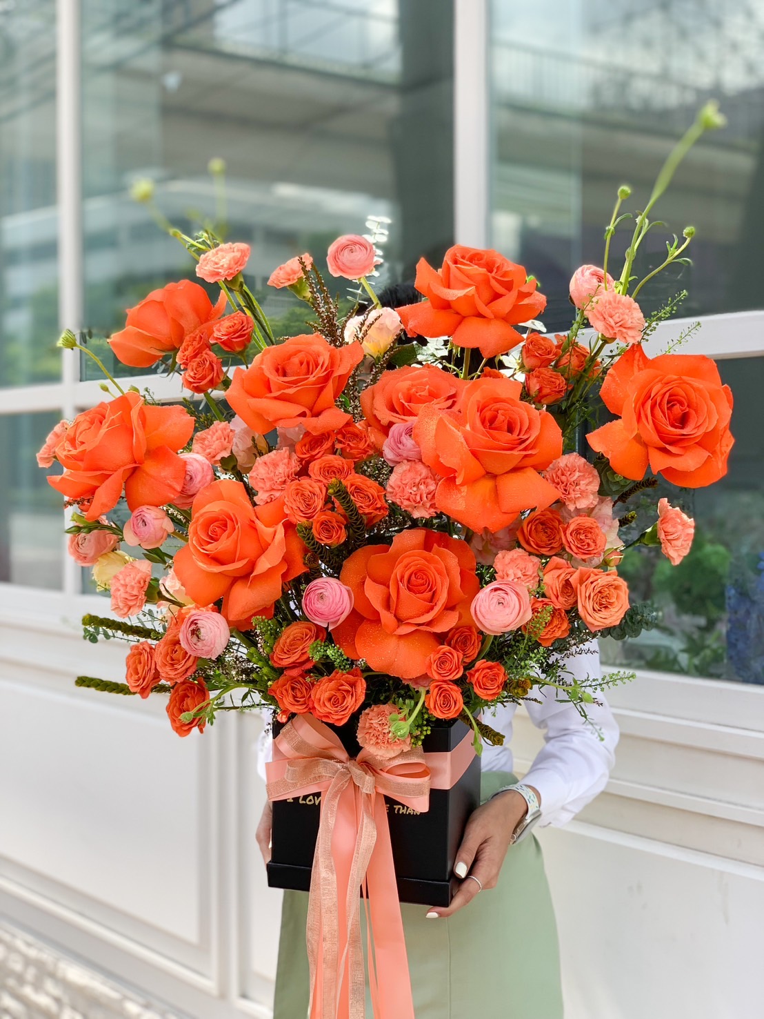 A person holding a large, vibrant bouquet consisting of orange and pink roses with greenery from Scarlet Blooms. The romantic arrangement is wrapped in black paper and adorned with a peach-colored ribbon. The background features a modern building with large windows.