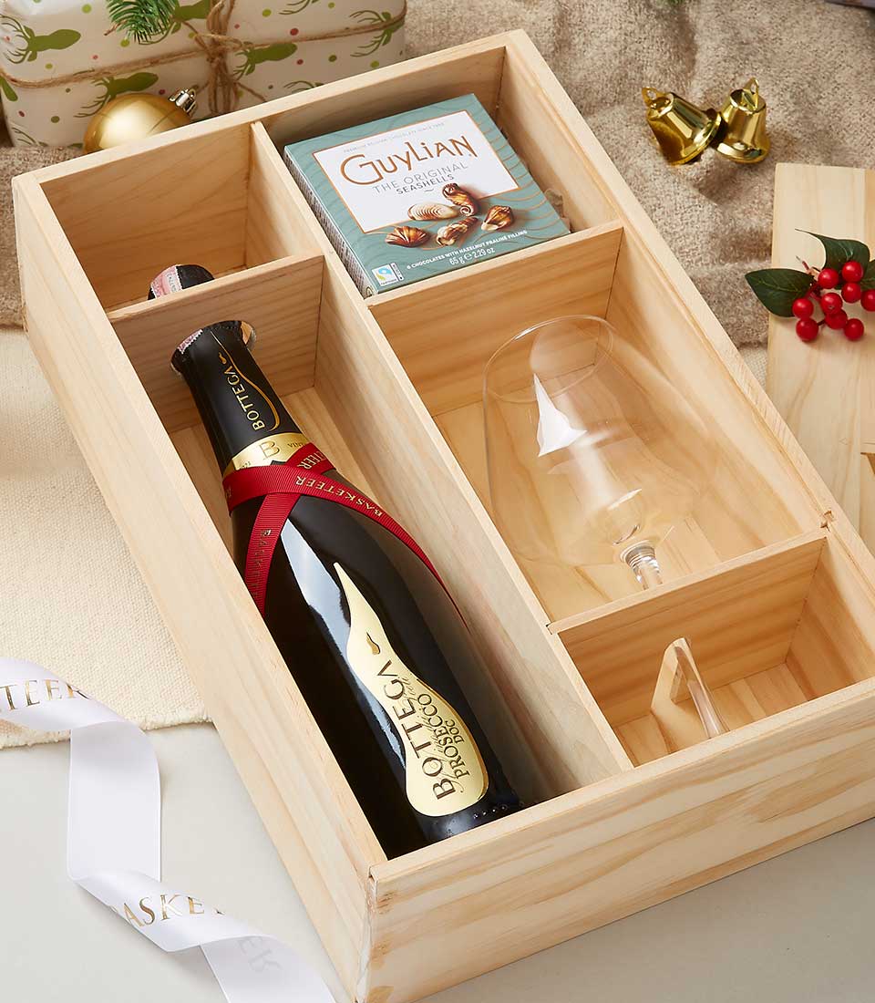 A beautifully crafted wooden gift box showcasing a bottle of luxurious Bottega sparkling wine, an elegantly packaged box of Guylian Belgian chocolates, and a pristine empty wine glass. Adorned with festive decorations including shimmering ornaments and vibrant holly, the scene is further elevated by a glimpse of a white ribbon accented with gold text from Basketeer.