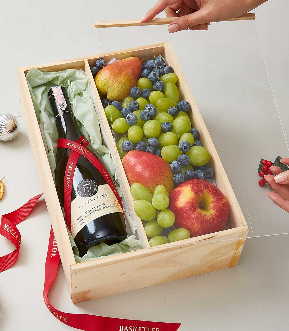 A lavish wooden gift box from Basketeer, elegantly designed to delight the senses, contains a meticulously wrapped bottle of exquisite red wine. Next to it, an assortment of crisp apples, succulent pears, verdant green grapes, and plump blueberries lies invitingly. An individual is gracefully placing a clear cover over this bountiful arrangement. A luxurious red ribbon adorned with the esteemed 