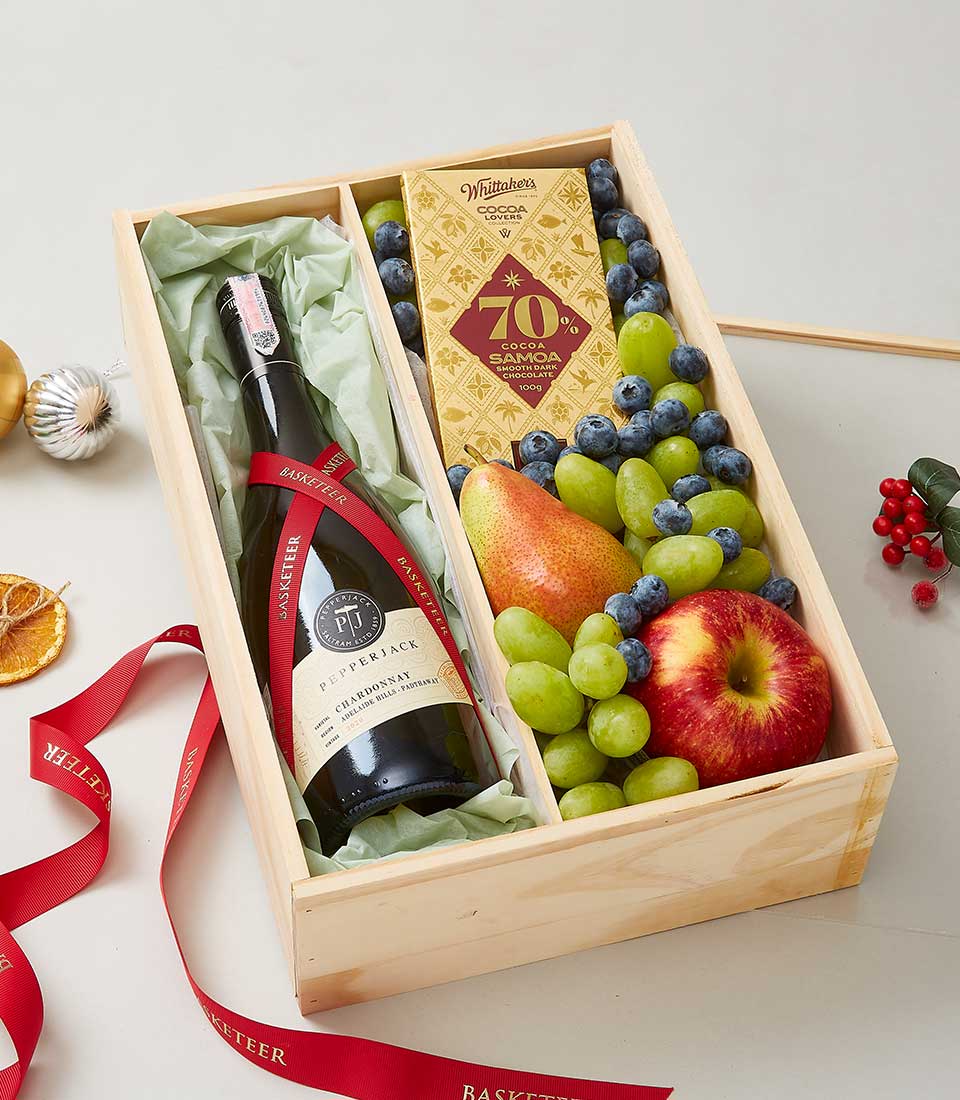 An elegant rectangular wooden gift box from Basketeer featuring a fine bottle of Pepperjack wine, a rich bar of chocolate, fresh green grapes, plump blueberries, and a crisp red apple. The box is adorned with a luxurious red ribbon emblazoned with the Basketeer logo. Surrounding the scene are tasteful Christmas decorations, enhancing the festive and inviting atmosphere.