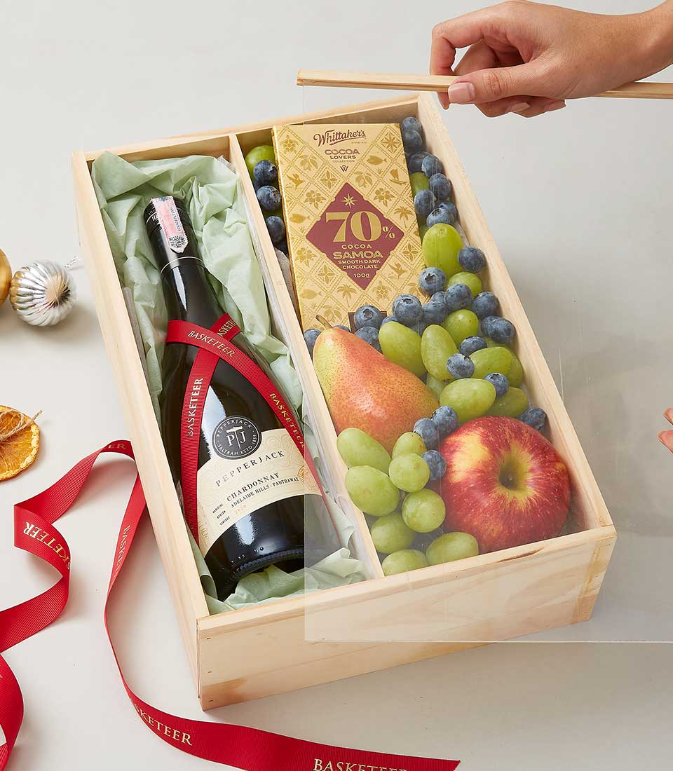 A beautifully crafted wooden gift box, elegantly adorned with the Basketeer logo, reveals a sumptuous collection tailored for luxury indulgence. Nestled inside is a sophisticated bottle of Pepperjack Shiraz wine, an artisanal bar of 70% dark chocolate, and a vibrant array of fresh fruits including crisp apples, luscious green grapes, and plump blueberries. Accents of decorative red ribbons add a festive touch. The scene captures the moment where a hand delicately lifts the clear cover from this opulent bounty, inviting you to savor the exquisite offerings from Basketeer.