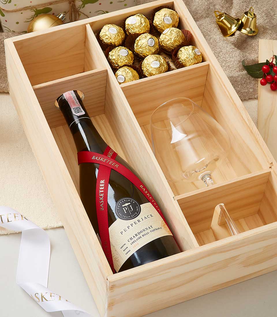 A wooden gift box containing a bottle of Pepperjack wine, a wine glass, and several gold-wrapped chocolates in a compartment. The box is positioned on a beige surface, with a white ribbon and festive decorations nearby.