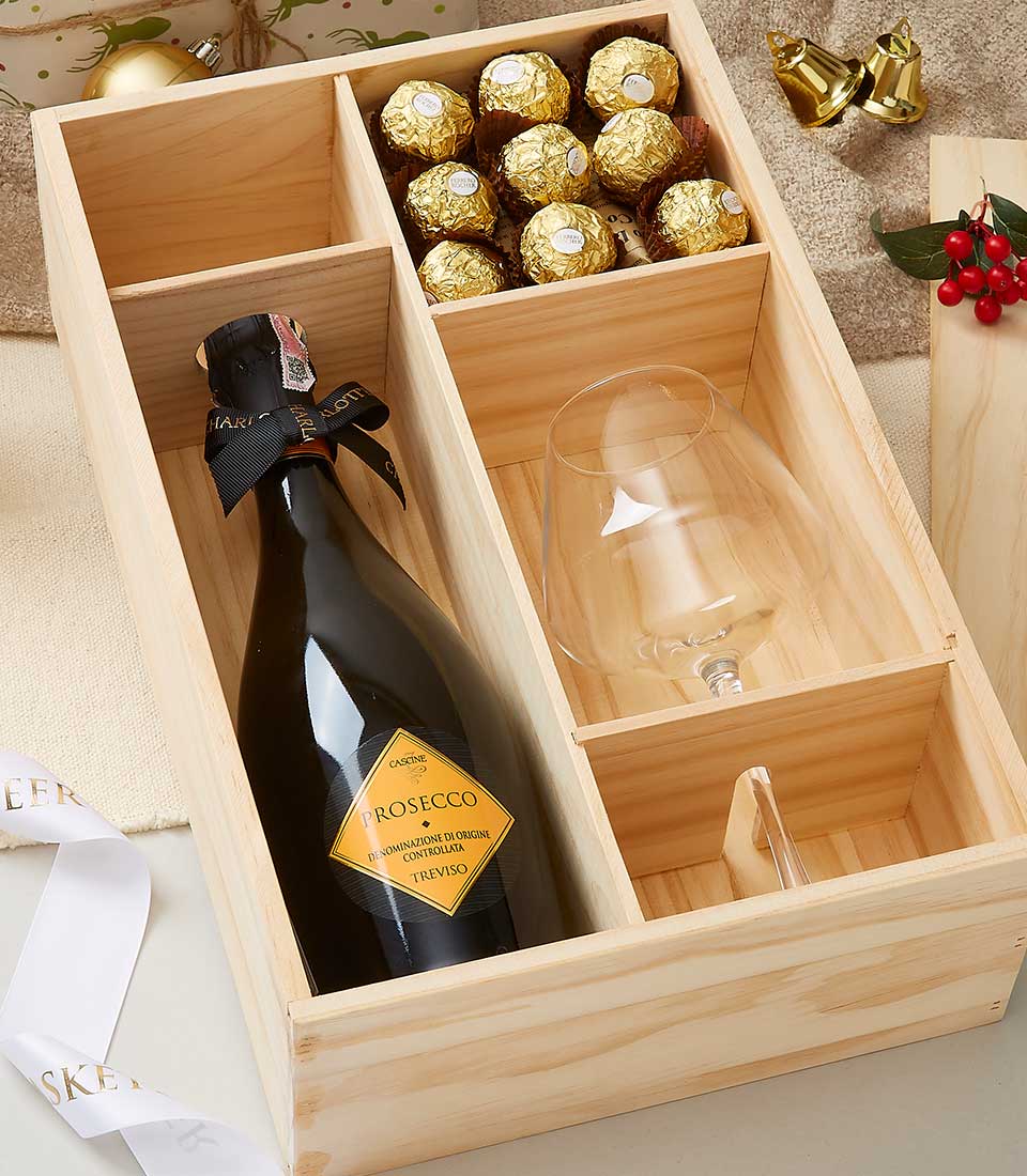 A lavish wooden gift box showcases a bottle of premium prosecco, accompanied by a solitary, exquisitely crafted wine glass. Nestled alongside are eight Ferrero Rocher chocolates, elegantly wrapped in shimmering gold foil. A festive holly sprig accentuates the luxurious presentation. The prestigious Basketeer branding adorns the surrounding white and gold ribbons and accompanying packaging, all set gracefully against a soft beige backdrop.
