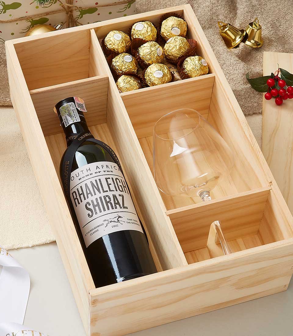 A wooden gift box containing a bottle of South African Rhanleigh Shiraz wine, a wine glass, and a section with Ferrero Rocher chocolates. The box is surrounded by festive decorations, including golden jingle bells and holly with red berries.
