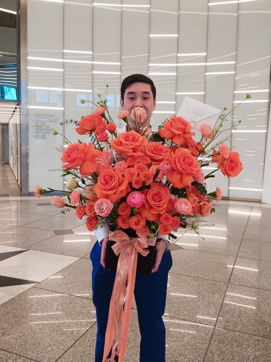 A person in a white shirt and blue pants stands indoors holding a large bouquet of orange and pink scarlet blooms with a white card attached. The background features a modern interior with glossy flooring and wall panels, evoking an air of romance.