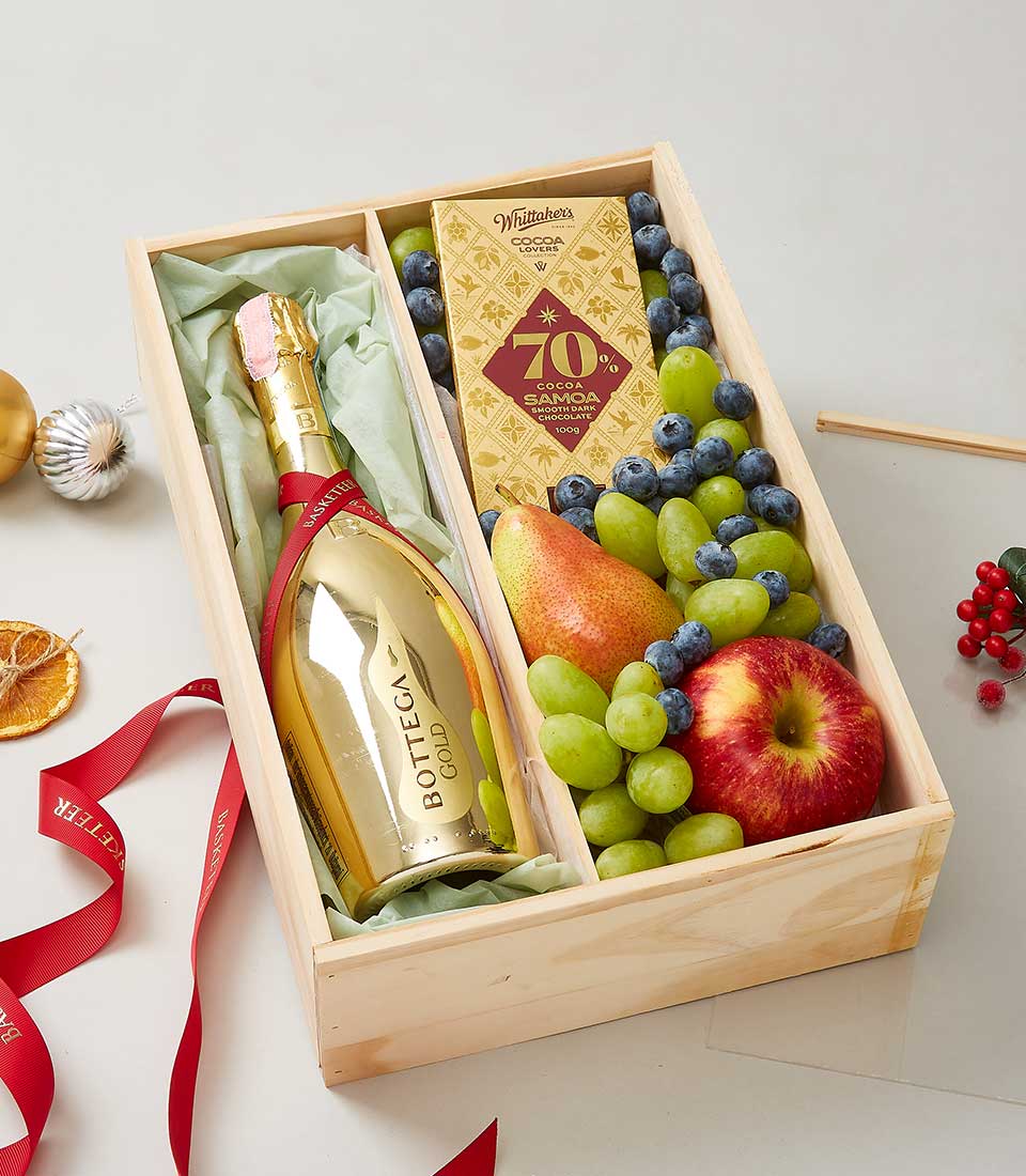 An elegant wooden gift box from Basketeer showcases sophistication with a bottle of Bottega Gold champagne nestled inside. Complementing this are an exquisite bar of Whittaker's 70% cocoa dark chocolate and a selection of fresh, vibrant fruits, including red and green apples, plump grapes, and luscious blueberries. The luxurious presentation is enhanced by a rich red ribbon adorned with the Basketeer logo and tasteful festoon elements, making it an inviting sight that promises indulgence.
