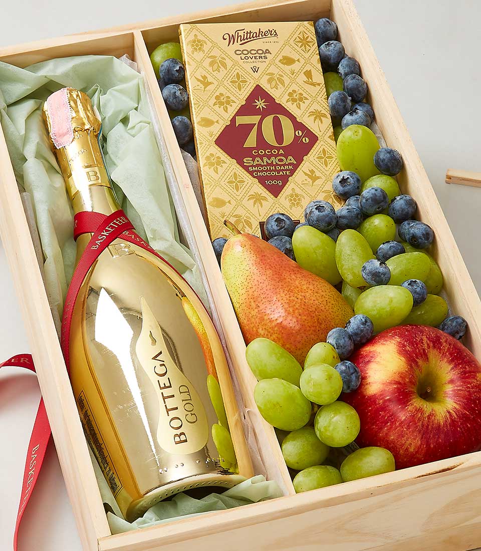 A luxurious wooden gift box from Basketeer, elegantly arranged to delight the senses. At its heart lies a stunning gold bottle of Bottega Gold sparkling wine, glistening with opulence. Accompanying this indulgence is a bar of rich Whittaker's 70% Cocoa Samoa dark chocolate. Fresh green grapes, plump blueberries, a crisp red apple, and a perfectly ripe pear are carefully nestled amidst verdant green tissue paper, creating an inviting scene of sophistication and taste-ready to be revealed at the lift of the Basketeer-branded ribbon or box lid.