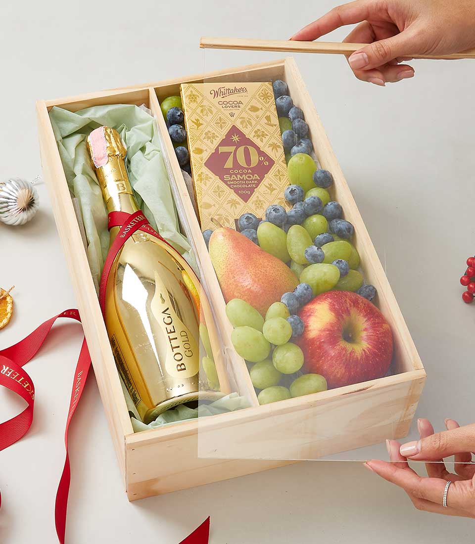 Alt text: An exquisite wooden gift box from Basketeer adorned with a delicate branded ribbon showcases a luxurious arrangement. Inside, you'll find a lavish gold bottle of Bottega Gold sparkling wine, an artisanal bar of Villars 70% cocoa chocolate, and an assortment of ripe fruits including lush green grapes, plump blueberries, and crisp apples. Two hands carefully place the elegant lid bearing the Basketeer logo onto the box, sealing its opulent contents within.