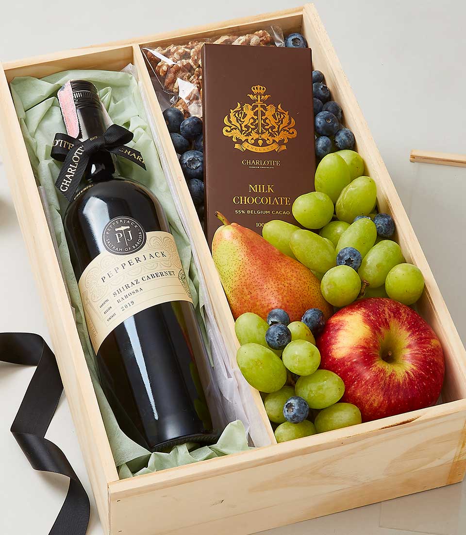 A luxurious wooden gift box by Basketeer, exquisitely curated to delight the senses. Nestled within is a bottle of fine red wine, artfully positioned on a bed of crinkled, golden paper. Surrounding this centerpiece are an array of delectable treasures: a silky bar of milk chocolate, a perfectly ripe pear, and a crisp apple. Juxtaposed hues of plump green and blue grapes add vibrancy, while an assortment of premium nuts provides the finishing touch. The scene exudes opulence and invitation, with touches of elegance signified by Basketeer's branded ribbon gracefully draped across the arrangement.