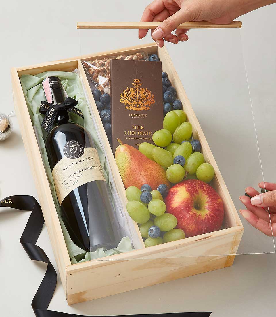 A lavish wooden gift box from Basketeer showcases an array of indulgent treats, including a bottle of exquisite red wine, a rich bar of milk chocolate, fresh green grapes, succulent blueberries, a crisp apple, and premium cashew nuts. A person gracefully lifts the clear plastic cover to reveal these delights. The hallmark black ribbon adorned with the Basketeer logo lies elegantly on the table beside the box.