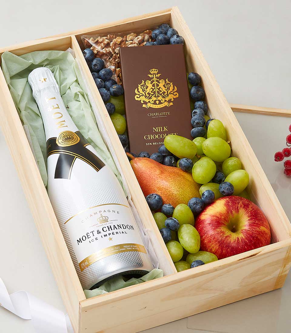 A luxurious wooden gift box elegantly presents a bottle of Moët & Chandon champagne, paired with a decadent chocolate bar in rich brown and gold wrapping. Nestled alongside are fresh, juicy apple and pear, a vibrant bunch of green grapes, succulent blueberries, and a curated selection of premium mixed nuts. An exquisite ribbon adorned with the Basketeer logo gracefully completes this opulent ensemble.