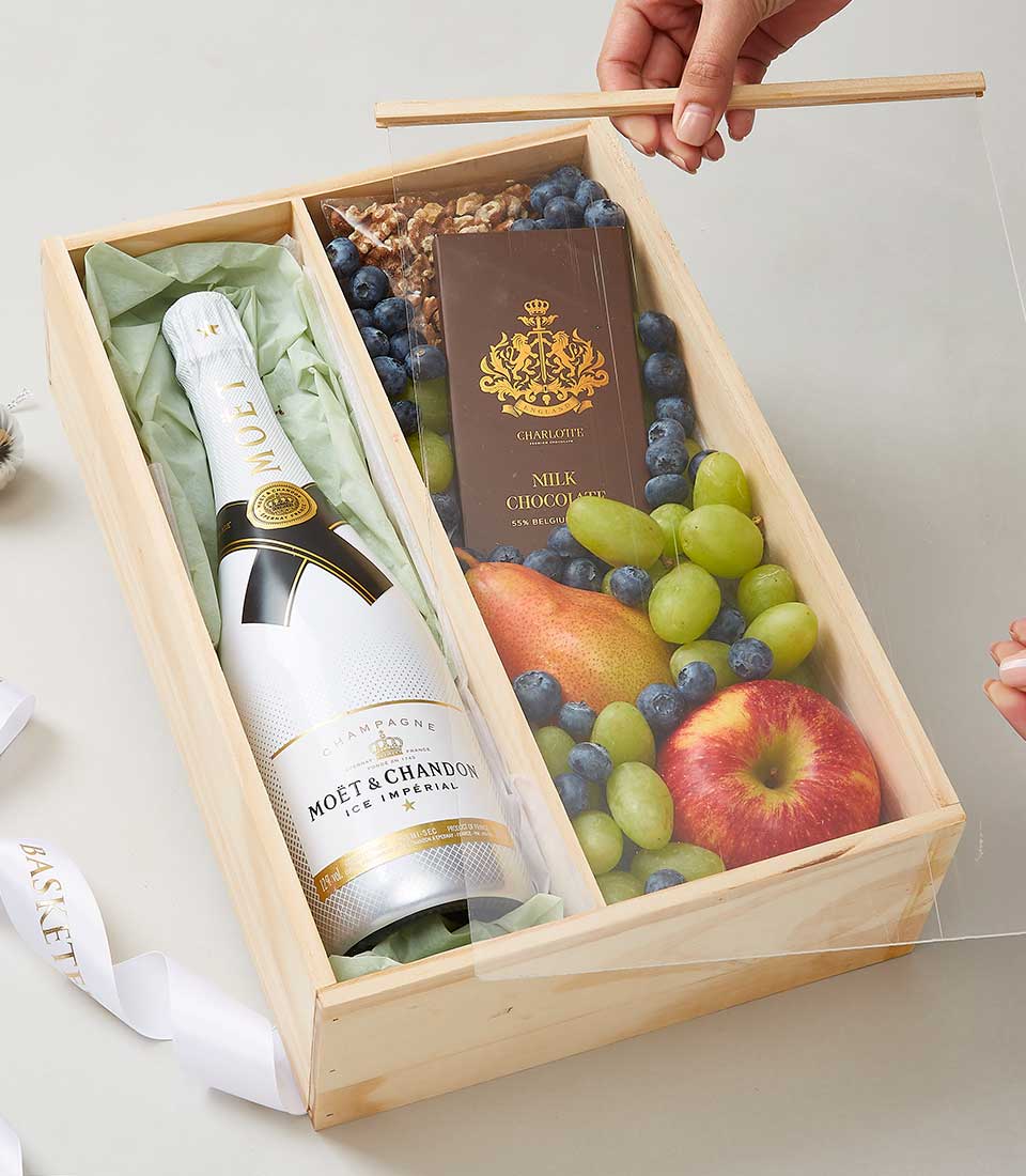 A luxurious wooden gift box from Basketeer, elegantly designed and brimming with indulgent delights. The left compartment cradles a bottle of Moët & Chandon Ice Imperial Champagne, promising a celebration in every sip. The right compartment showcases an exquisite array of fresh green grapes, juicy blueberries, a crisp apple, a succulent pear, a decadent chocolate bar and an assortment of gourmet mixed nuts. A person gracefully places the clear cover over this opulent ensemble, ensuring every item remains pristine within its Basketeer-branded confines—an invitation to pure luxury.
