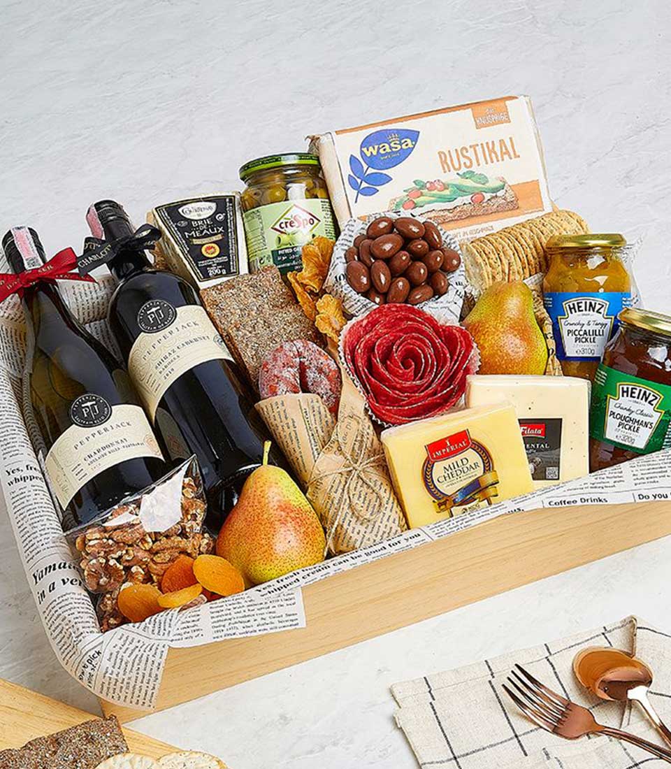 A gourmet gift basket with two bottles of red wine, assorted cheeses, pears, nuts, chocolates, and gourmet snacks like crackers and jars of mustard and pickles. The items are neatly arranged in a wooden tray lined with decorative paper. A cheese knife and cloth napkin are nearby for the perfect wine pair.