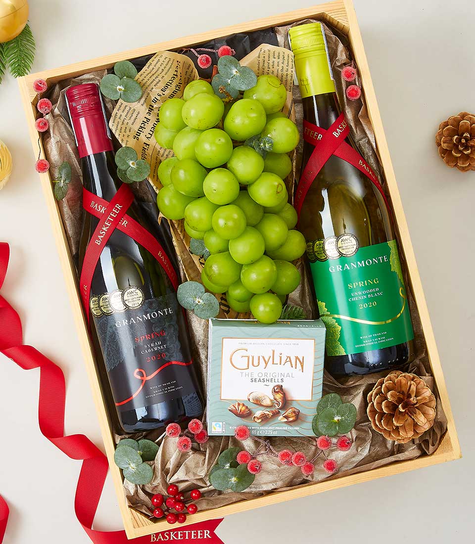 A luxurious wooden gift box adorned with the Basketeer logo, meticulously arranged to captivate. Inside, you'll find two exquisite bottles of Grammonte wine—one rich red and one crisp white—paired with a sumptuous box of Guylian Belgian chocolates. Nestled beside a fresh bunch of green grapes, the arrangement is tastefully decorated with elegant pinecones, vibrant red berries, and a festive ribbon that exudes an inviting holiday charm. This opulent presentation promises to delight and impress at any occasion.