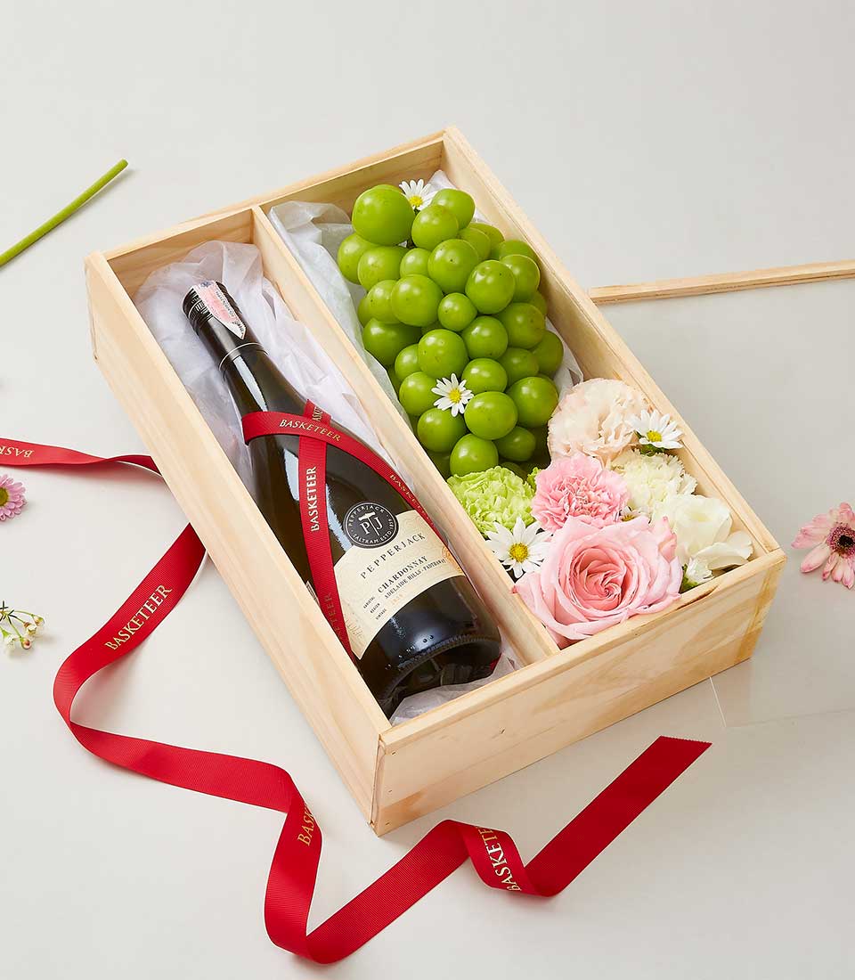 A luxurious wooden gift box by Basketeer showcases an elegant bottle of red wine nestled on the left side, accompanied by a lavish assortment of fresh green grapes, delicate pink roses, and exquisite white and pink blooms on the right. The scene is accentuated by a refined red ribbon adorned with the 