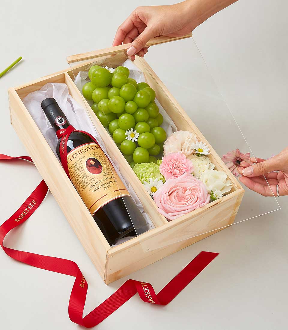 A luxurious wooden gift box by Basketeer, exquisitely designed to captivate. On one side, a fine bottle of wine awaits indulgence, while the other cradles a lush selection of fresh green grapes and an enchanting array of pink and white roses. A person with graceful hands is gently placing a transparent lid on this elegant ensemble. The scene is accentuated by a rich red ribbon adorned with the Basketeer logo, lying gracefully beside the box, completing this invitation to opulence.