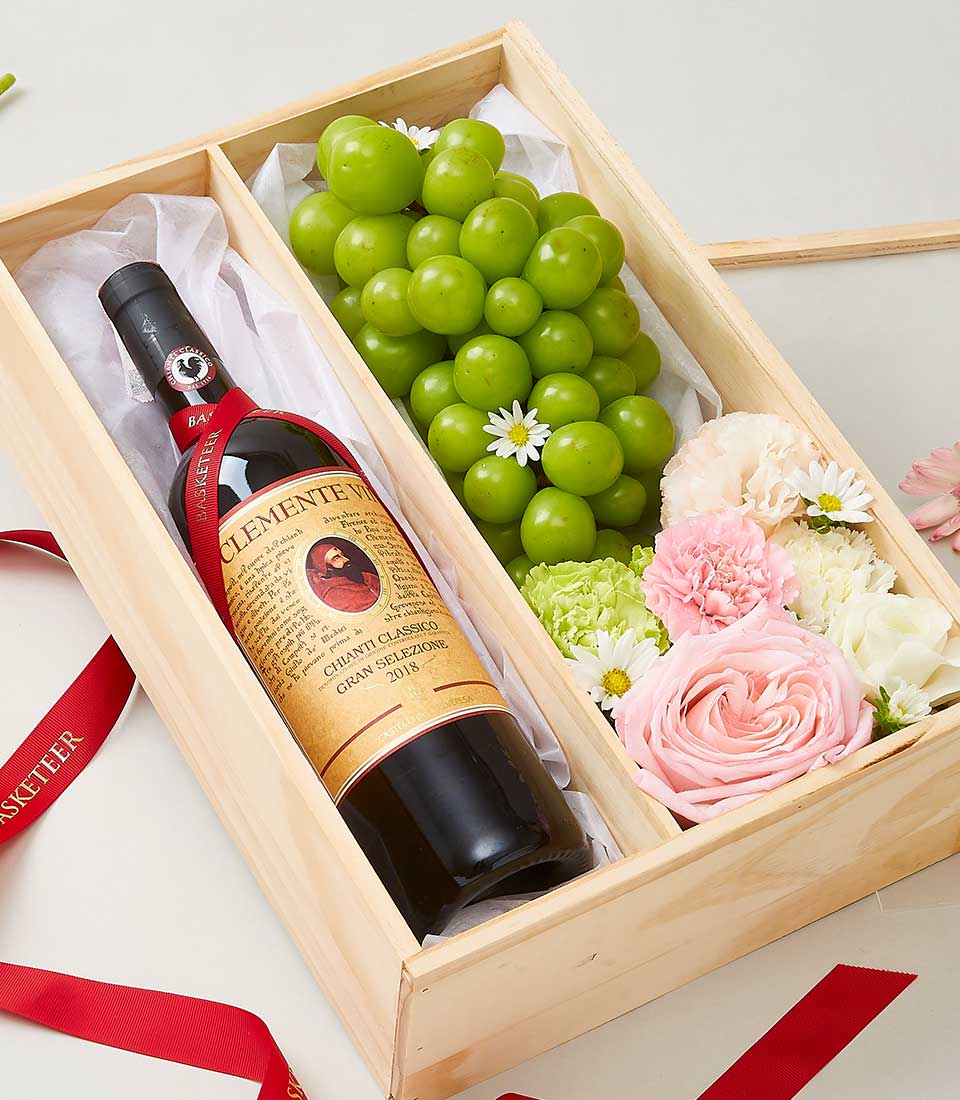 A luxurious wooden Basketeer gift box, elegantly adorned with a silky red ribbon, houses a fine bottle of red wine. Beside it lies a vibrant medley of fresh green grapes, complemented by an opulent bouquet of mixed flowers featuring delicate pink roses, pristine white daisies, and charming carnations. Exquisite red ribbons cascade gracefully around the open Basketeer box, creating an inviting and sumptuous scene.