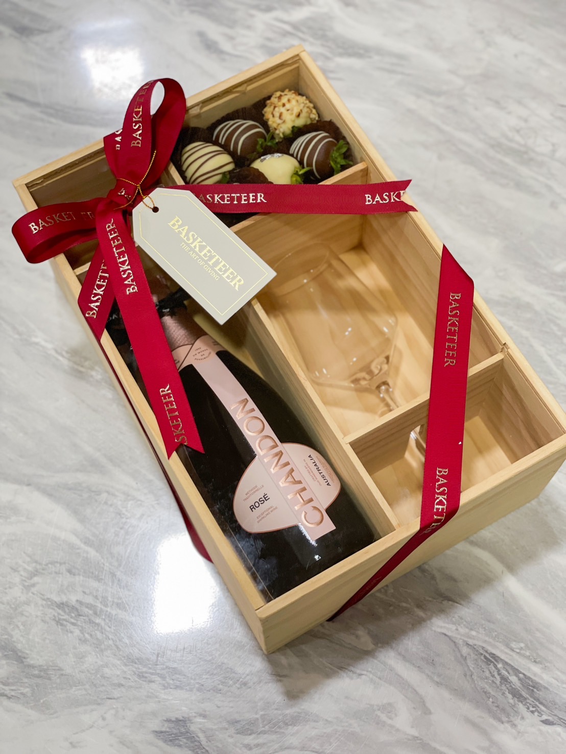 Wooden gift box with a premium bottle, gourmet chocolates, and elegant glassware, tied with a red ribbon by Basketeer.