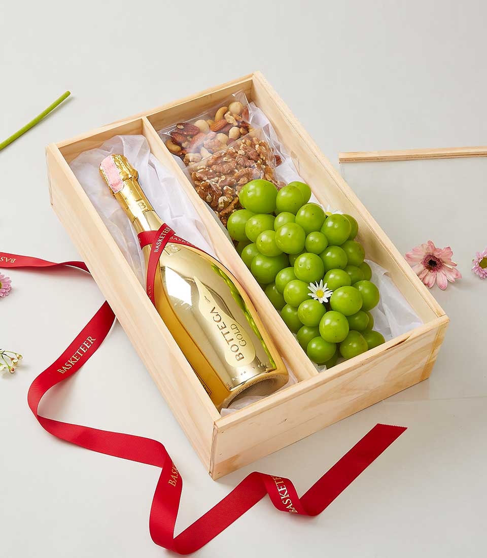 A luxurious wooden gift box by Basketeer showcases an exquisite bottle of Bottega Gold Prosecco, accented with a distinctive pink neck seal. Nestled beside the bottle, a cluster of vibrant green grapes adorned with an elegant white blossom adds a touch of freshness, while an assortment of premium mixed nuts invites indulgence. The box is elegantly tied with a Basketeer-branded red ribbon, exuding sophistication and care. Surrounding the box are scattered delicate pink flowers and lush green stems, enhancing the scene's inviting ambiance.