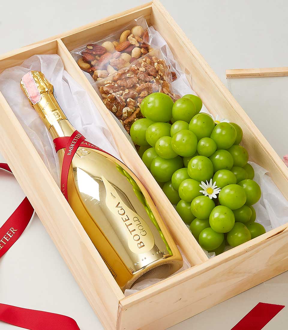 A luxurious wooden gift box featuring an elegant gold bottle of Bottega Gold Prosecco, nestled alongside a cluster of fresh green grapes adorned with a delicate flower, and an assortment of gourmet mixed nuts. The box is lined with pristine white tissue paper, and opulent red ribbons from Basketeer add a touch of sophistication.