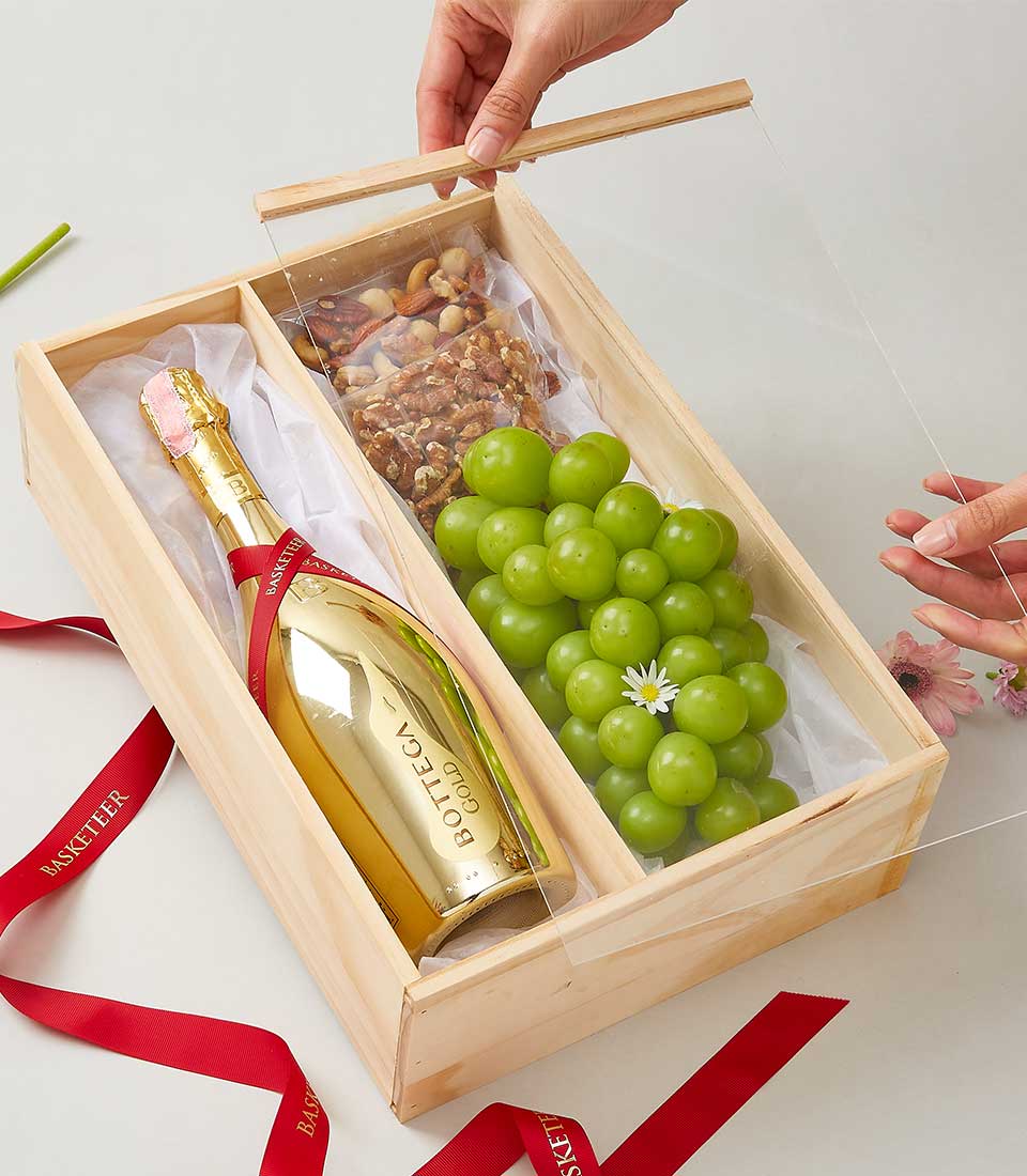 A person is gracefully unveiling an exquisite wooden gift box, elegantly adorned with the distinguished Basketeer logo. Nestled within, a luxurious bottle of Bottega Gold sparkling wine gleams invitingly. Accompanying this indulgence is a vibrant bunch of green grapes, crowned with a delicate flower, and an opulent assortment of mixed nuts. The contents sit atop pristine white tissue paper for an added touch of refinement. A lavish red ribbon lies artfully beside the box, completing the scene with a hint of sumptuous allure.