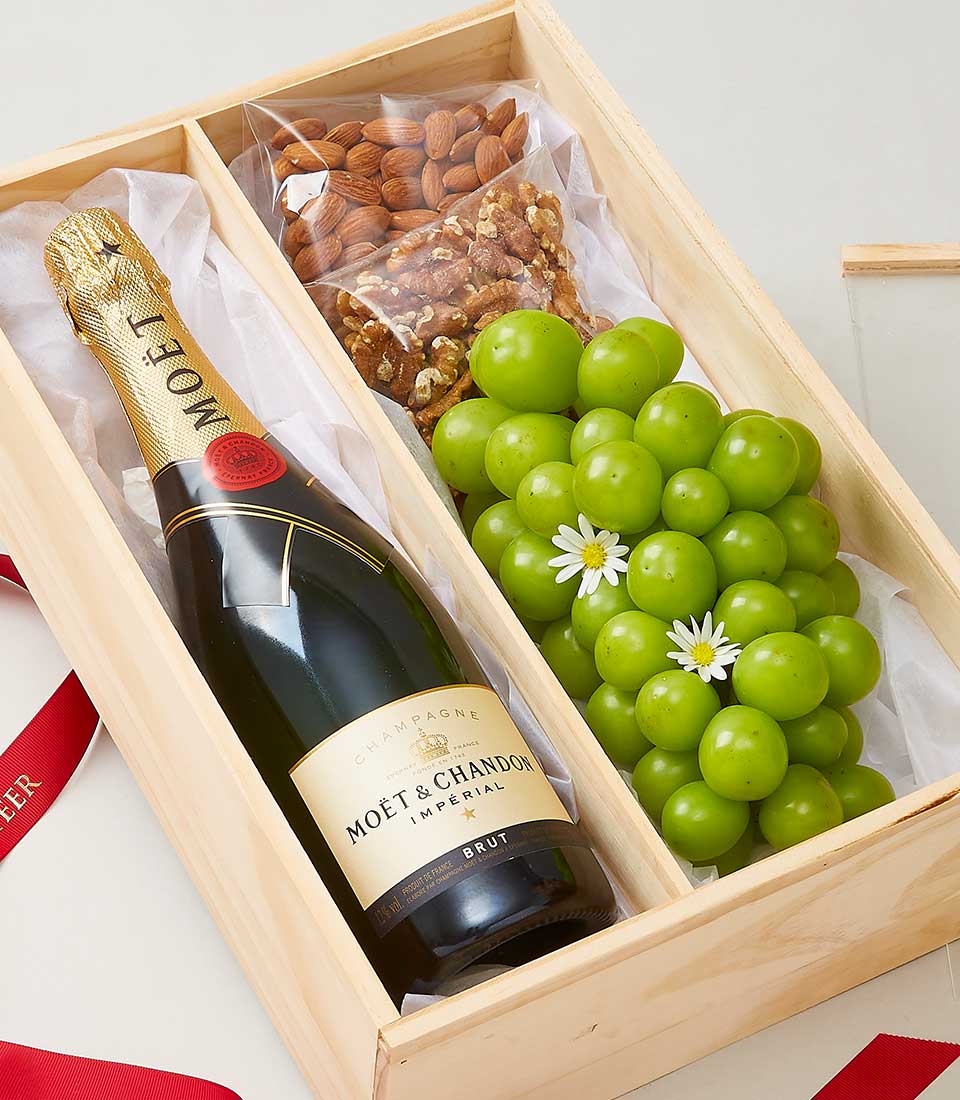 A meticulously crafted wooden gift box from Basketeer, elegantly lined with pristine white tissue paper. Inside, a prestigious bottle of Moët & Chandon champagne promises opulence and delight. Accompanying this indulgent experience is a selection of premium almonds and walnuts, offering rich flavor and texture. A fresh bunch of luscious green grapes adorned with delicate small white flowers adds an inviting touch of nature’s elegance. In the background, the scene is gracefully accentuated by a luxurious red ribbon signifying warmth and celebration.