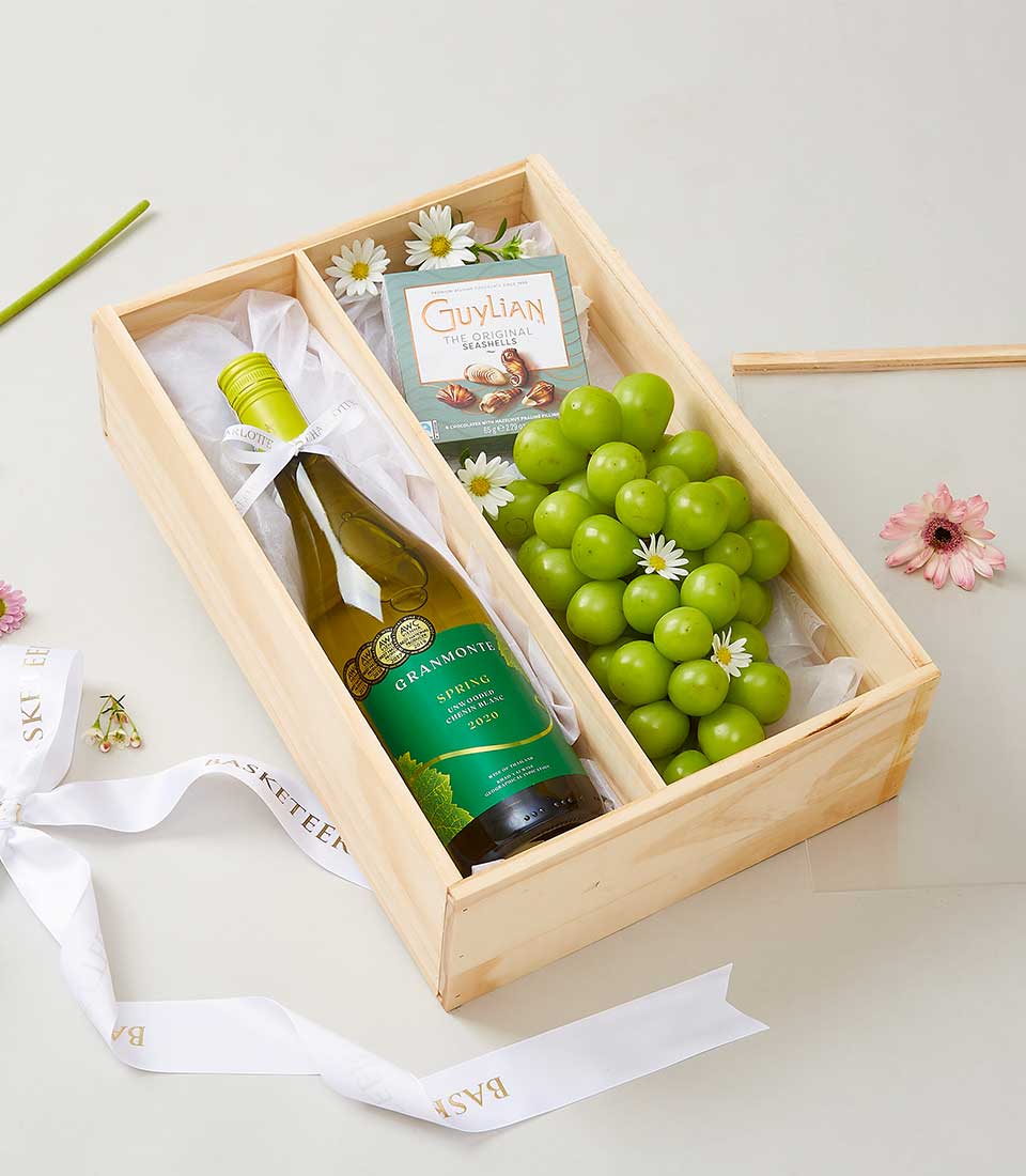 A luxurious wooden gift box adorned with an elegant white ribbon from Basketeer, brimming with opulence. Inside, you'll discover a bottle of the exquisite Grant Burge wine, a succulent bunch of green grapes, and an indulgent box of Guylian chocolates. The scene is further enriched by the delicate scatterings of small white and pink daisies, offering an inviting touch to this refined presentation.