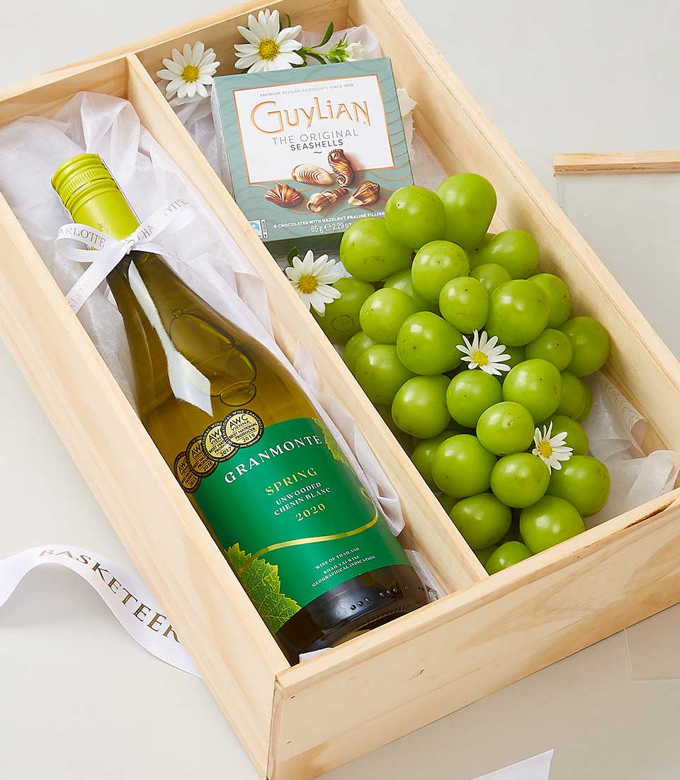A luxurious wooden gift box adorned with the prestigious Basketeer logo, containing an exquisite bottle of Grammonte white wine, a bountiful bunch of fresh green grapes delicately intertwined with elegant white flowers, and an indulgent box of Guylian chocolate seashells. The interior is lined with sumptuous white fabric and embellished with a sophisticated white ribbon featuring 