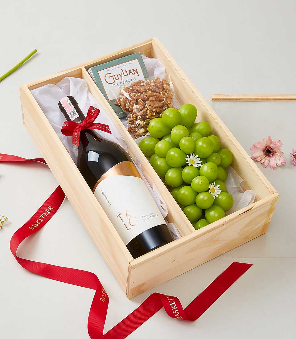 A luxurious wooden gift box from Basketeer elegantly presents an exquisite bottle of wine adorned with a red Basketeer ribbon, a refined small box of Guylian chocolates, a fresh bundle of green grapes, and a premium bag of walnuts. The entire ensemble is tastefully enhanced with Basketeer-branded red ribbons. In the background, a delicate pink flower and an accompanying ribbon set the scene for this sophisticated and inviting presentation.