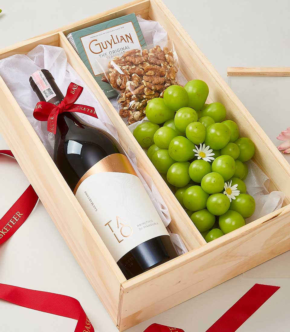 A luxurious wooden gift box, branded with the Basketeer logo, cradles an exquisite bottle of red wine wrapped elegantly in a lush red ribbon. Nestled alongside are a cluster of vibrant green grapes, delicately adorned with charming white flowers. Adding to the opulence is a pack of decadent Guylian chocolate-covered nuts. The interior is partially lined with crisp white tissue paper, while an inviting red ribbon lies gracefully nearby.