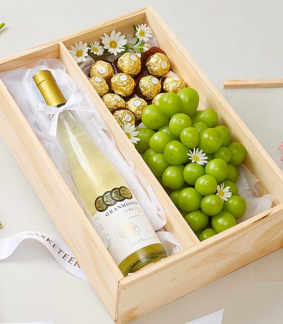 An exquisite wooden gift box showcasing a prestigious bottle of white wine, artfully adorned with a delicate white ribbon bearing the Basketeer logo. Nestled beside are luxurious Ferrero Rocher chocolates and a fresh, vibrant bunch of green grapes, elegantly accented with dainty white flowers. Each item is meticulously arranged on soft tissue paper, epitomizing the refined elegance and thoughtful presentation synonymous with Basketeer.