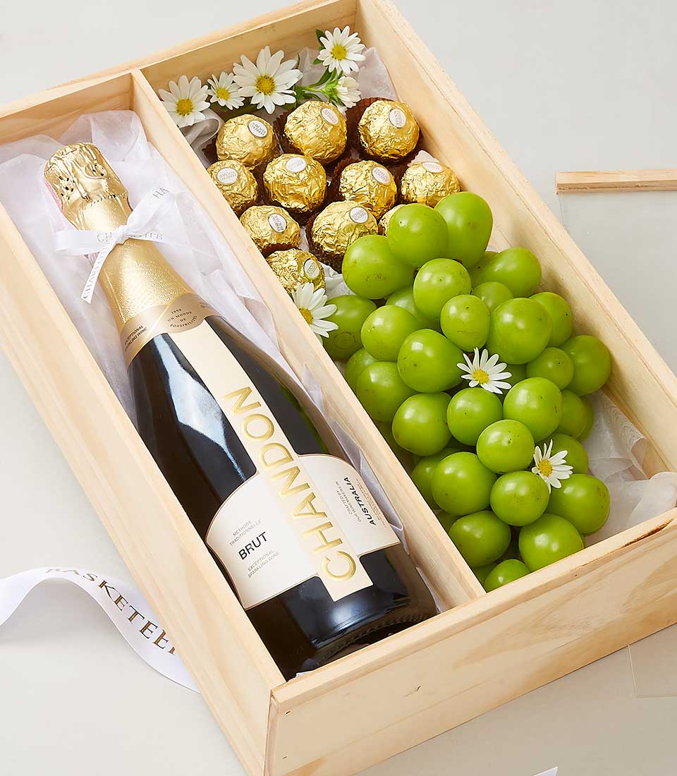 A tastefully curated wooden gift box from Basketeer, featuring a bottle of luxurious Chandon Brut champagne. Inside, fresh green grapes and indulgent Ferrero Rocher chocolates are elegantly arranged alongside delicate small white flowers. The interior is lined with premium tissue paper, adding an extra touch of sophistication. Completing the presentation is a beautifully tied ribbon adorned with the distinguished 