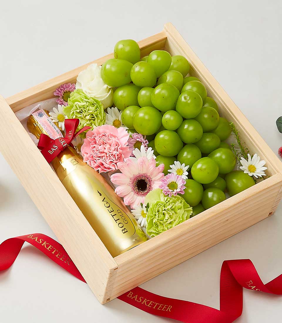 An elegant wooden gift box featuring a lush bottle of Bottega Gold Prosecco, accompanied by a vibrant bunch of fresh green grapes. Surrounding the opulent selections are exquisite pink and white floral blooms, interwoven with luxurious red ribbons adorned with the 
