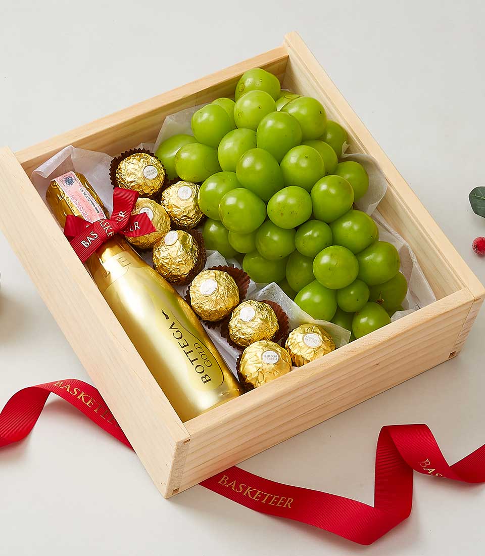 A luxurious wooden gift box elegantly cradles a selection of plump green grapes, an array of decadent Ferrero Rocher chocolates, and a petite bottle of exquisite Bottega Gold Prosecco. The sophisticated presentation is completed with a rich red ribbon inscribed with the distinguished 