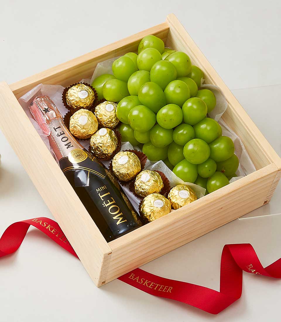 A lavish wooden box from Basketeer presents an exquisite ensemble featuring a vibrant bunch of green grapes, eleven decadent Ferrero Rocher chocolates, and a sophisticated mini bottle of Moët champagne. The box is elegantly open, revealing the indulgent treats within, while a luxurious red ribbon bearing the 