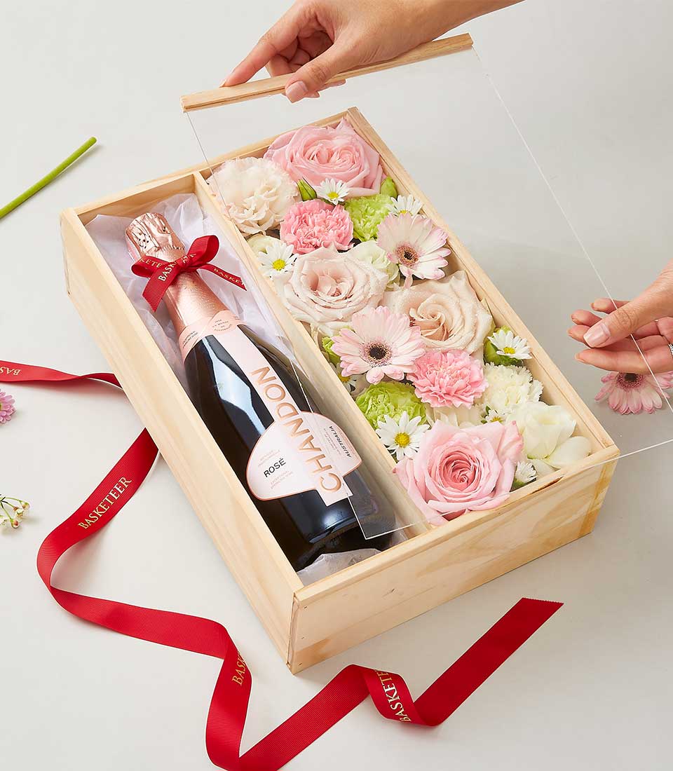 An exquisite wooden gift box by Basketeer, featuring a bottle of Chandon Rosé sparkling wine nestled amidst an elegant array of blush pink and pristine white flowers, including luxurious roses and delicate chrysanthemums. A pair of hands gracefully lifts the transparent lid, unveiling this opulent floral presentation. A sophisticated red ribbon bearing the word 